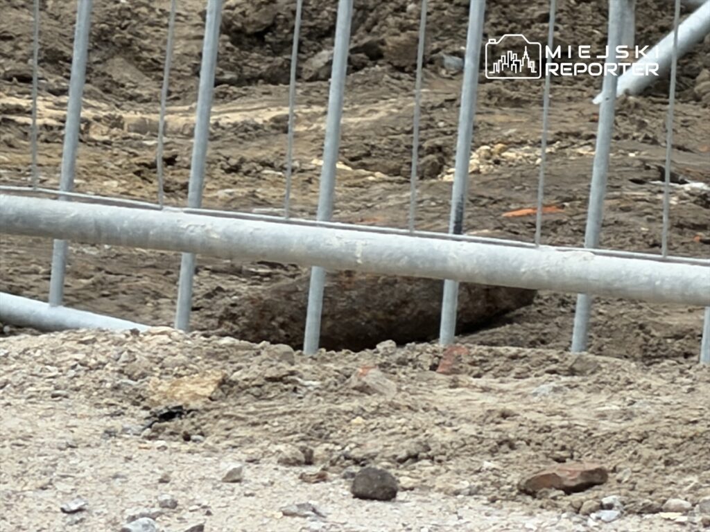 Wykop na placu budowy z metalowym ogrodzeniem, w tle widoczna ziemia i kamienie.