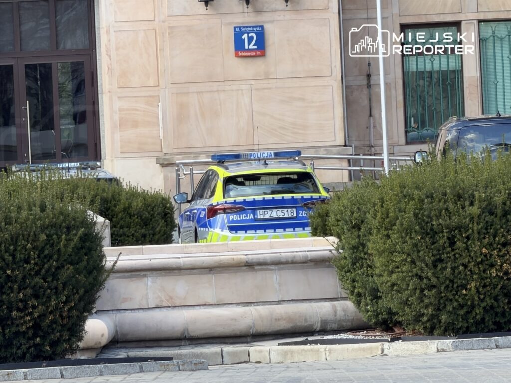 Policyjny radiowóz zaparkowany przed budynkiem z numerem 12, otoczony niskimi krzewami.