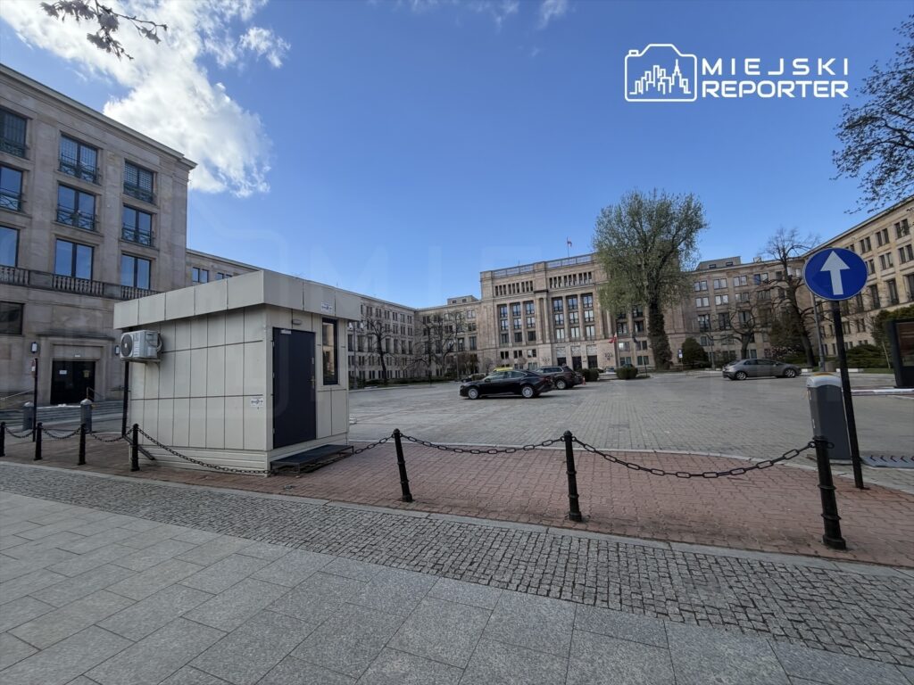 Na placu przed nowoczesnym budynkiem znajduje się stróżówka z klimatyzacją, otoczona łańcuchem.