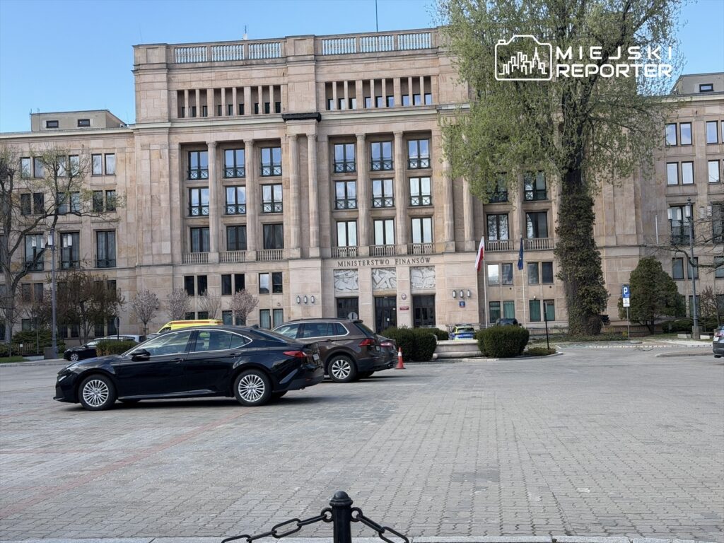 Czarne samochody zaparkowane przed budynkiem Ministerstwa Finansów z widocznymi flagami.
