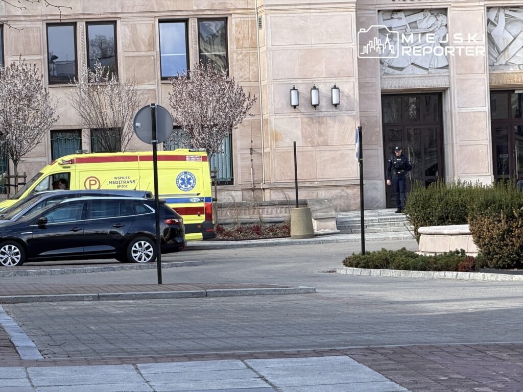 Karetka pogotowia z napisem „WSPIRAJ MEDITRANS” parkuje obok czarnego samochodu, a policjant stoi przy wejściu do budynku.