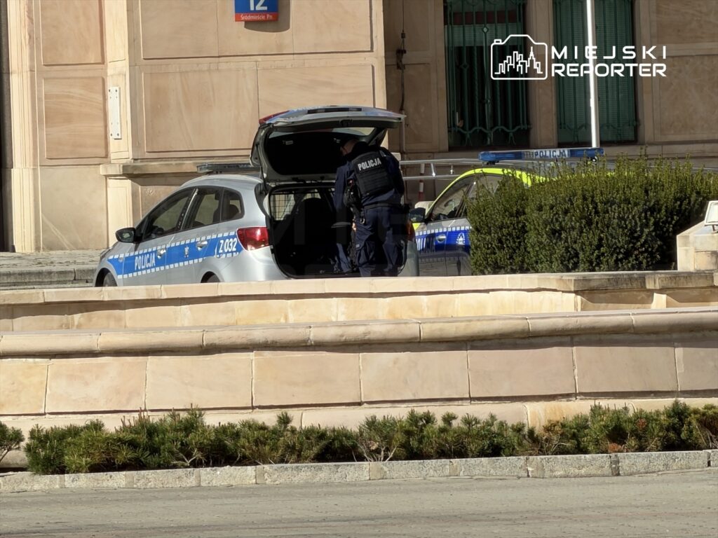 Policjant przeszukuje bagażnik radiowozu zaparkowanego obok drugiego pojazdu policyjnego.