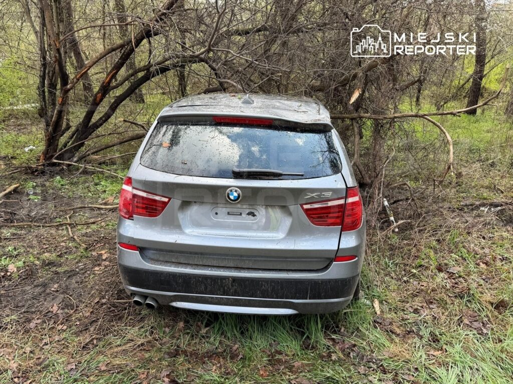 Zniszczony samochód marki BMW X3 zaparkowany w lesie, otoczony mokrą trawą i gałęziami drzew.