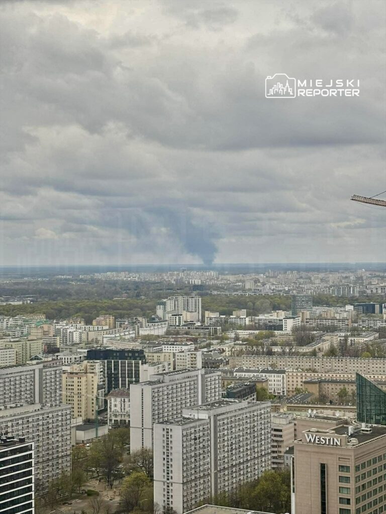 Widok na miejską panoramę z chmurami w tle, w oddali widoczny dym nad zabudowaniami.