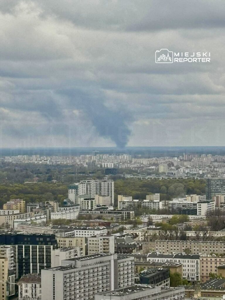 Ciemny dym unoszący się nad miastem w tle, w otoczeniu bloków mieszkalnych i zieleni.