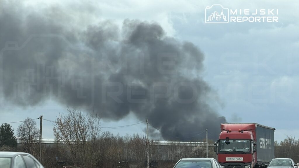 Czarny dym unoszący się z miejsca pożaru w tle, z ciężarówką jadącą w kierunku kamery na drodze.