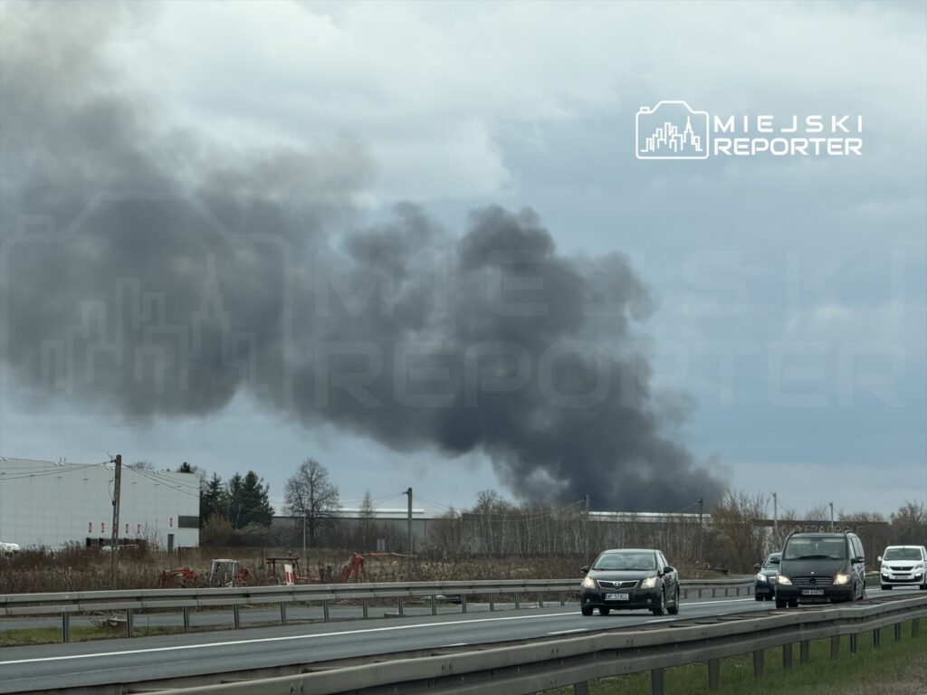 Czarny dym unoszący się nad terenami przemysłowymi, w tle widoczne samochody na drodze.