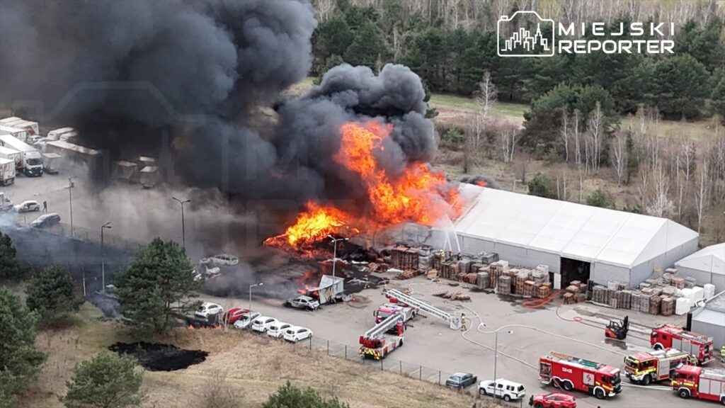 Wielka pożar w magazynie, ogień i dym unoszą się nad terenem, a w pobliżu działania straży pożarnej.