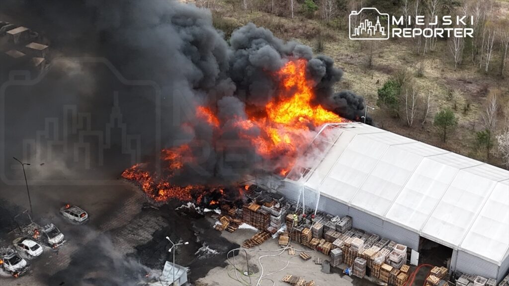 Pożar w magazynie z intensywnym dymem i płomieniami, w pobliżu widoczne zniszczone samochody i stosy drewnianych palet.