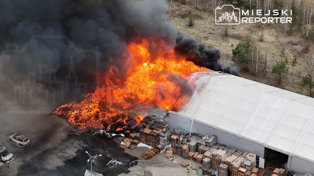 Pożar obejmujący dach magazynu, z intensywnym ogniem i gęstym dymem, w otoczeniu drewnianych palet.