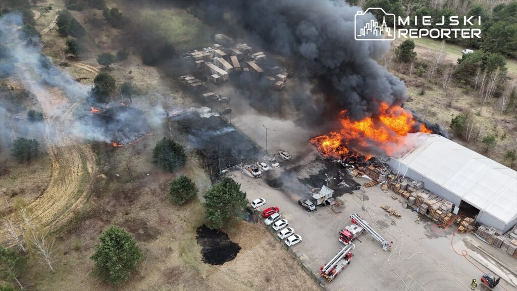 Pożar trawi magazyn i pobliskie tereny, dym i płomienie unoszą się w powietrzu, widać wozy strażackie.
