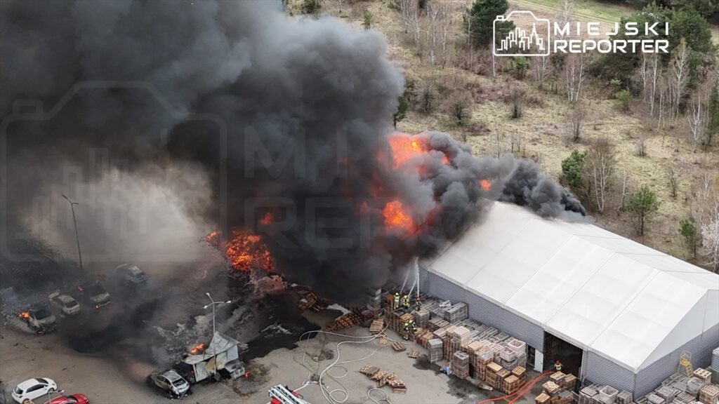 Pożar w magazynie, z którego wydobywa się gęsty, czarny dym oraz płomienie, obok strażacy gaszą ogień.