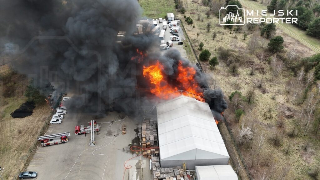 Ogromne płomienie wydobywające się z dachu magazynu, otoczone gęstym czarnym dymem, a w tle widać zaparkowane samochody i wóz strażacki.