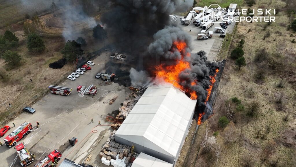 Pożar dużej hali przemysłowej, z której wydobywa się gęsty czarny dym i płomienie, w otoczeniu zaparkowanych samochodów i drzew.