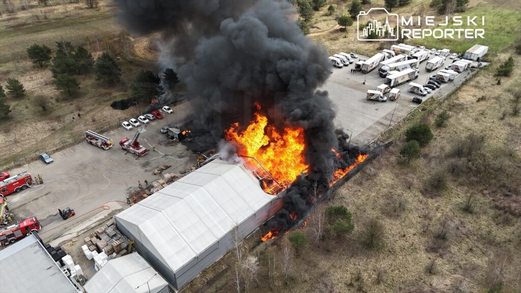 Pożar dużego budynku przemysłowego, z którego wydobywają się płomienie i gęsty dym. W tle widoczne wozu strażackie oraz osoby pracujące przy akcji gaśniczej.