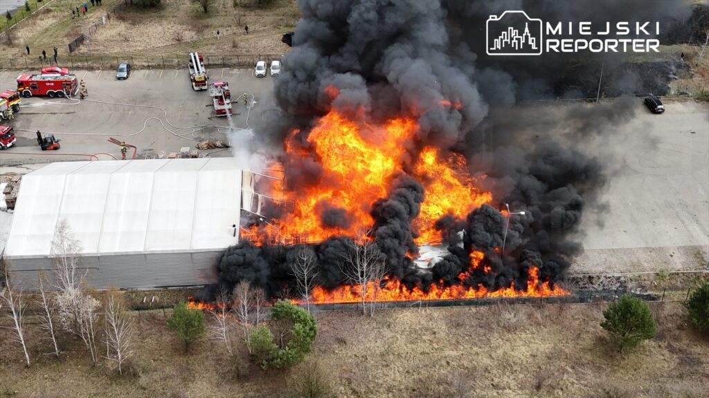 Ogromny pożar trawi halę magazynową, z której wydobywają się kłęby czarnego dymu i płomienie, w otoczeniu wozów strażackich.
