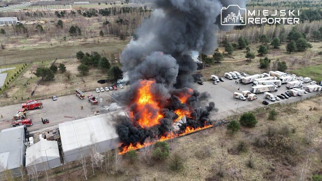Wysoka chmura czarnego dymu i ogień wydobywający się z budynku przemysłowego na terenie parkingu, w otoczeniu samochodów.