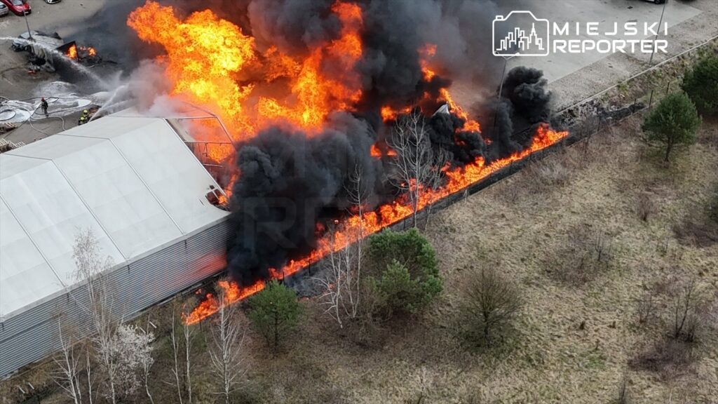Pożar budynku przemysłowego z intensywnymi płomieniami i czarnym dymem, w tle widoczni strażacy gaszący ogień.