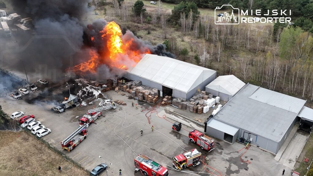 Wielki pożar na terenie zakładu z dymem i płomieniami wydobywającymi się z budynku; w pobliżu wozów strażackich.