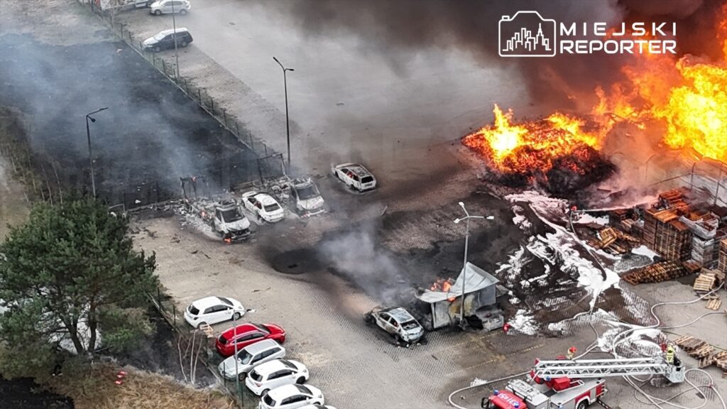 Płonące samochody i zniszczona budka na parkingu, obok widać dym i strażaków gaszących ogień.