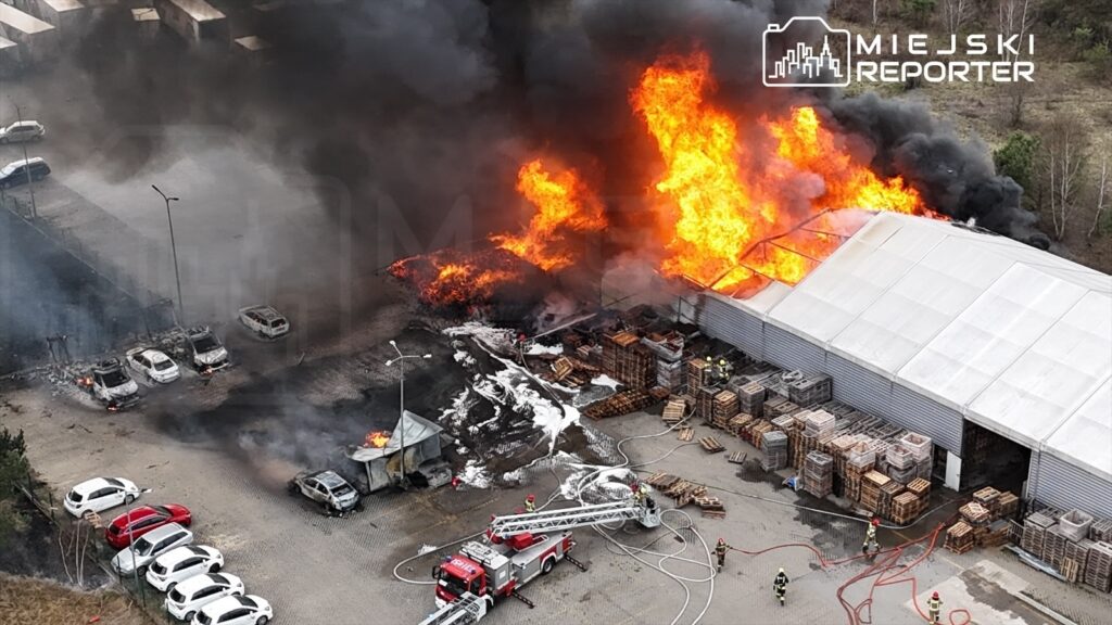 Ogień trawi halę magazynową, a strażacy gaszą pożar w otoczeniu zniszczonych pojazdów i palet.