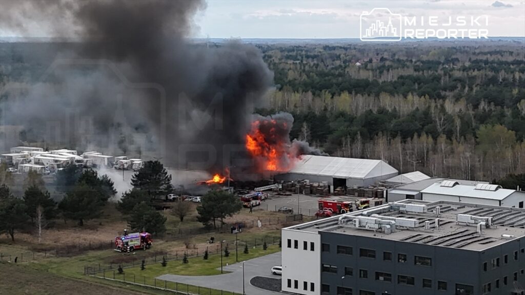Wielka chmura czarnego dymu unosząca się z płonącego budynku, w tle widoczne samochody strażackie i otaczająca zieleń.
