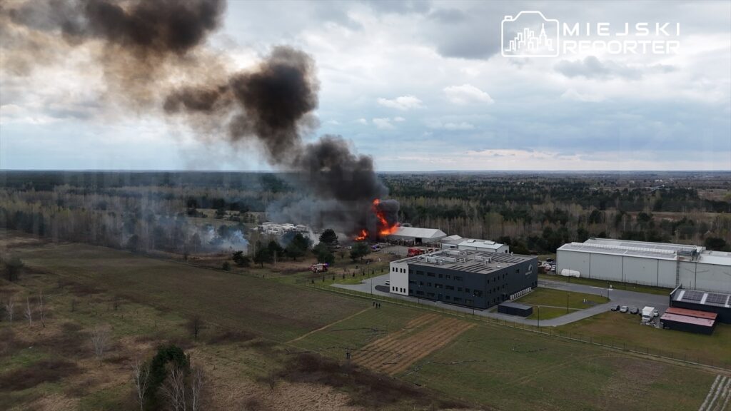 Pożar w zakładzie przemysłowym, z którego wydobywa się gęsty czarny dym, w otoczeniu lasu i pól.
