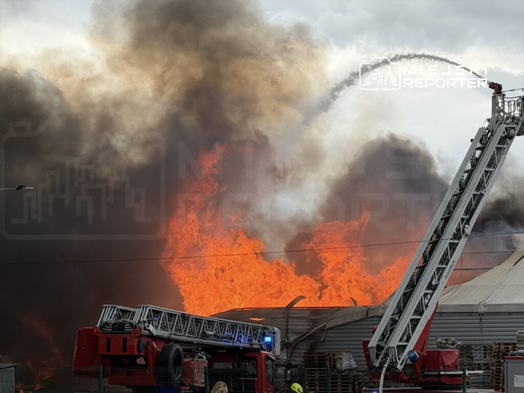 Pożar w magazynie, z płomieniami i gęstym dymem unoszącym się w powietrzu; w tle wóz strażacki z drabiną.