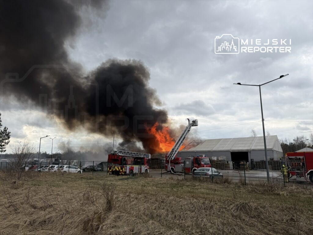 Wielka chmura czarnego dymu unosi się nad płonącym budynkiem, a w tle widać strażacki wóz z drabiną.