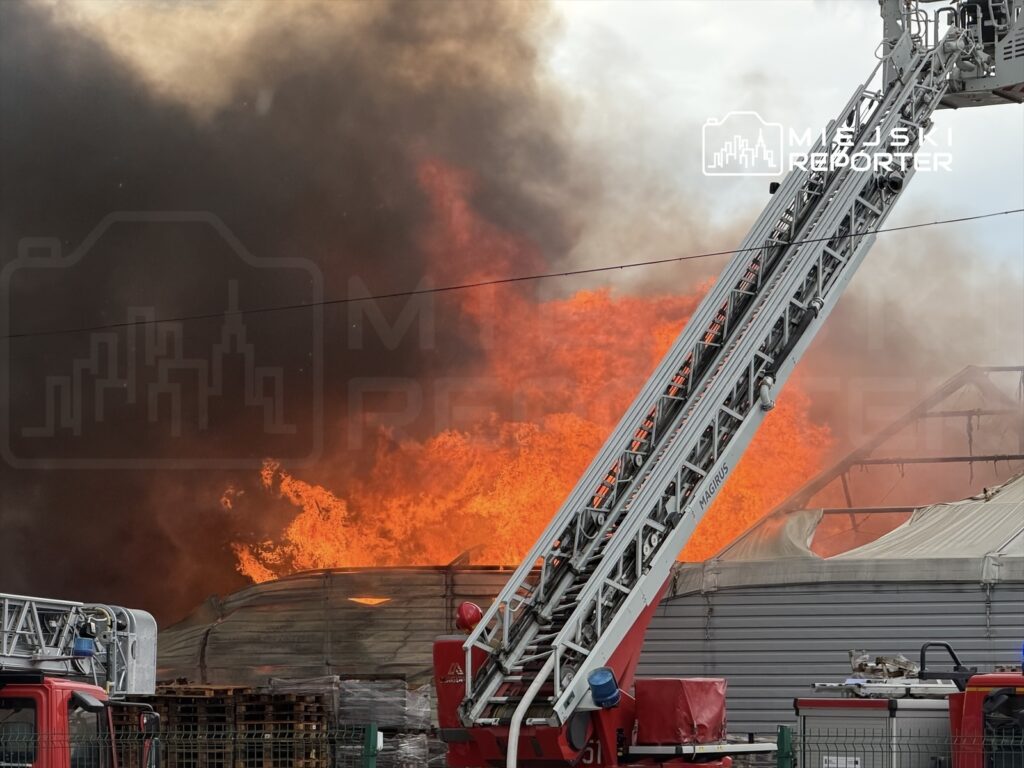 W tle ogień i dym wydobywający się z płonącego budynku, w pierwszym planie wóz strażacki z drabiną.