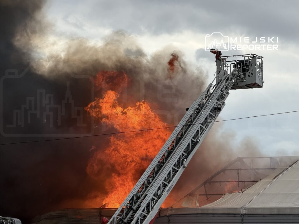 Strażak na podnośniku gaśniczym kieruje strumień wody na płonący budynek, otoczony gęstym dymem.