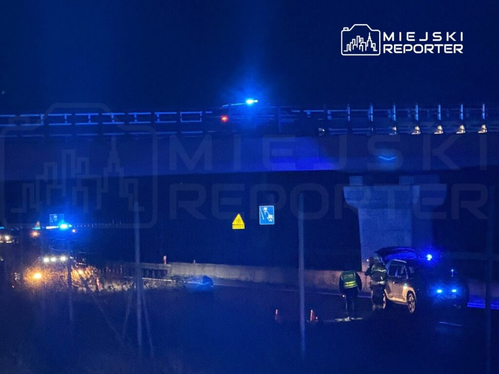 Na moście nocą, radiowozy z niebieskimi światłami zabezpieczają miejsce zdarzenia, przy którym pracują służby.