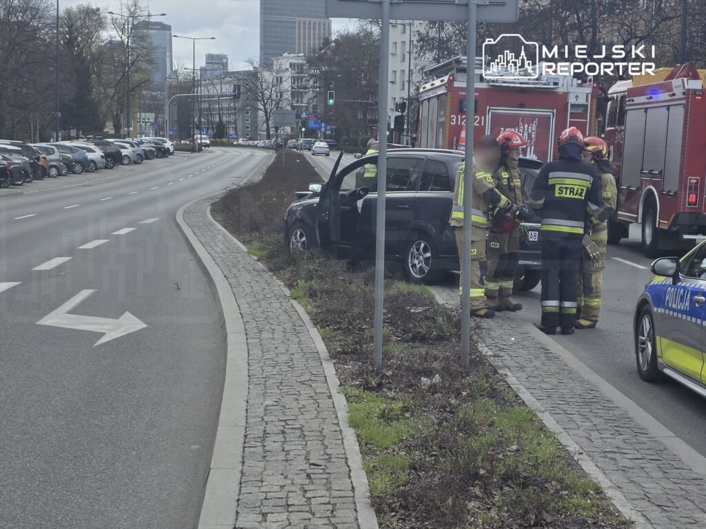 Grupa strażaków rozmawia obok uszkodzonego samochodu na ulicy, w tle widoczne pojazdy policyjne i strażackie.