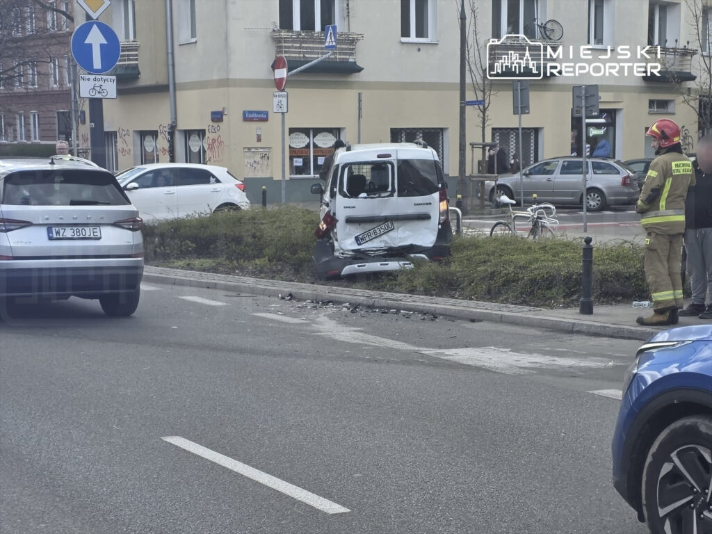 Zniszczony samochód dostawczy stał na poboczu drogi po kolizji, obok grupy osób i strażaka w mundurze.