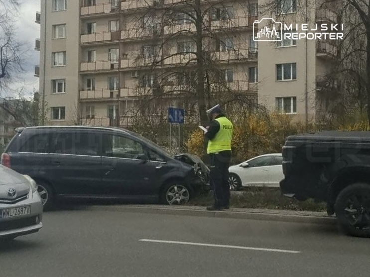 Policjant w kamizelce odblaskowej notuje zdarzenie przy uszkodzonym samochodzie na poboczu ruchliwej ulicy.