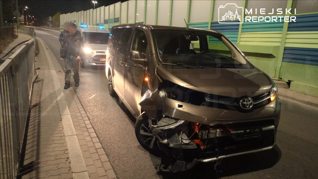 Mężczyzna w czarnej odzieży stoi obok uszkodzonego samochodu dostawczego na nocnej drodze, obok widać radiowóz.