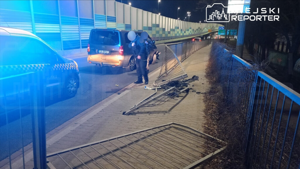 Funkcjonariusz policji bada uszkodzone ogrodzenie na chodniku obok drogi, gdzie stoi zaparkowany samochód.