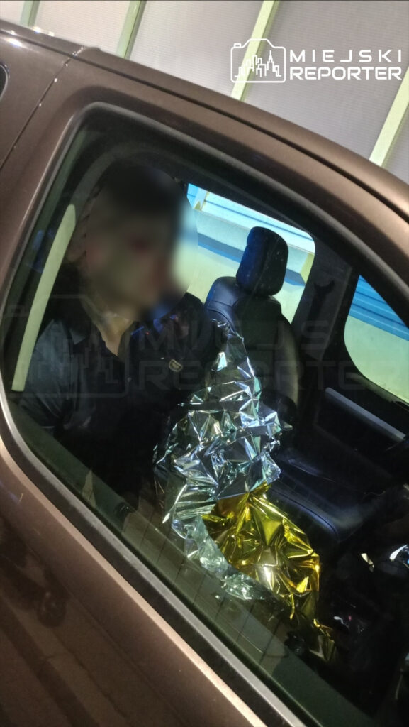 Mężczyzna w czarnej koszuli siedzący w samochodzie z foliowym kocem w dłoni. Wnętrze pojazdu o ciemnym wykończeniu.