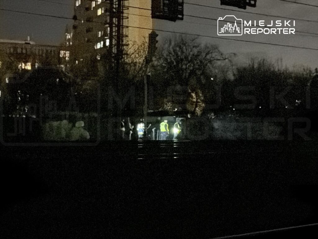 Grupa pracowników w kamizelkach odblaskowych prowadzi prace na torach kolejowych w ciemnej scenerii nocnej.