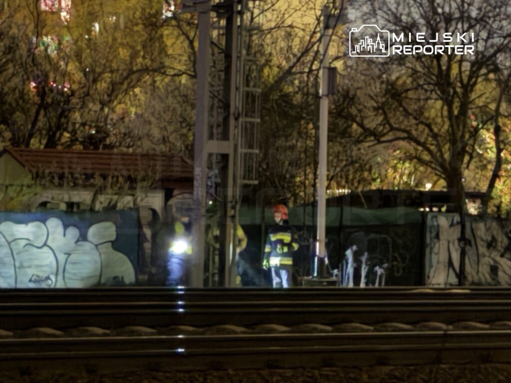 Dwóch pracowników w odblaskowych kamizelkach sprawdza torowisko w nocy, w tle widoczne graffiti na ogrodzeniu.