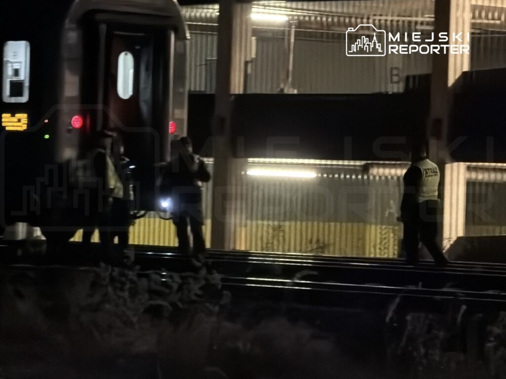 Grupa pracowników technicznych z latarkami sprawdzająca tory kolejowe w nocy obok pociągu na stacji.