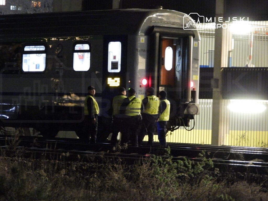 Grupa osób w odblaskowych kamizelkach rozmawia obok pociągu na stacji kolejowej w nocy.