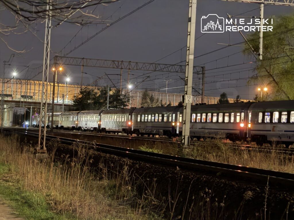 Pociąg osobowy zatrzymany na torach w nocy, w tle oświetlony budynek i linie energetyczne.