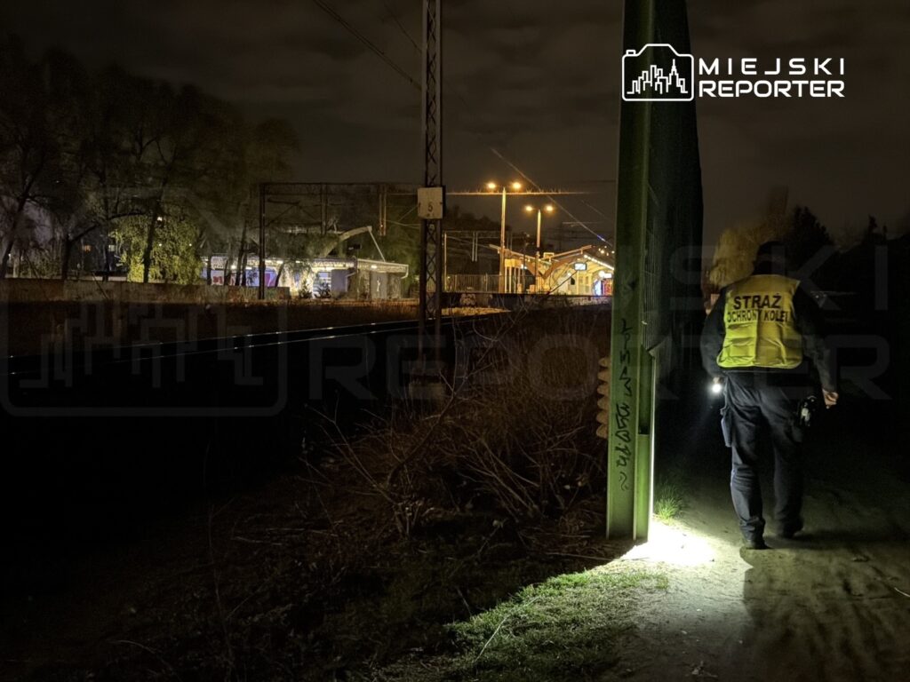 Strażnik kolejowy z latarką patroluje teren w pobliżu torów kolejowych w nocy, w tle widoczny budynek stacji.