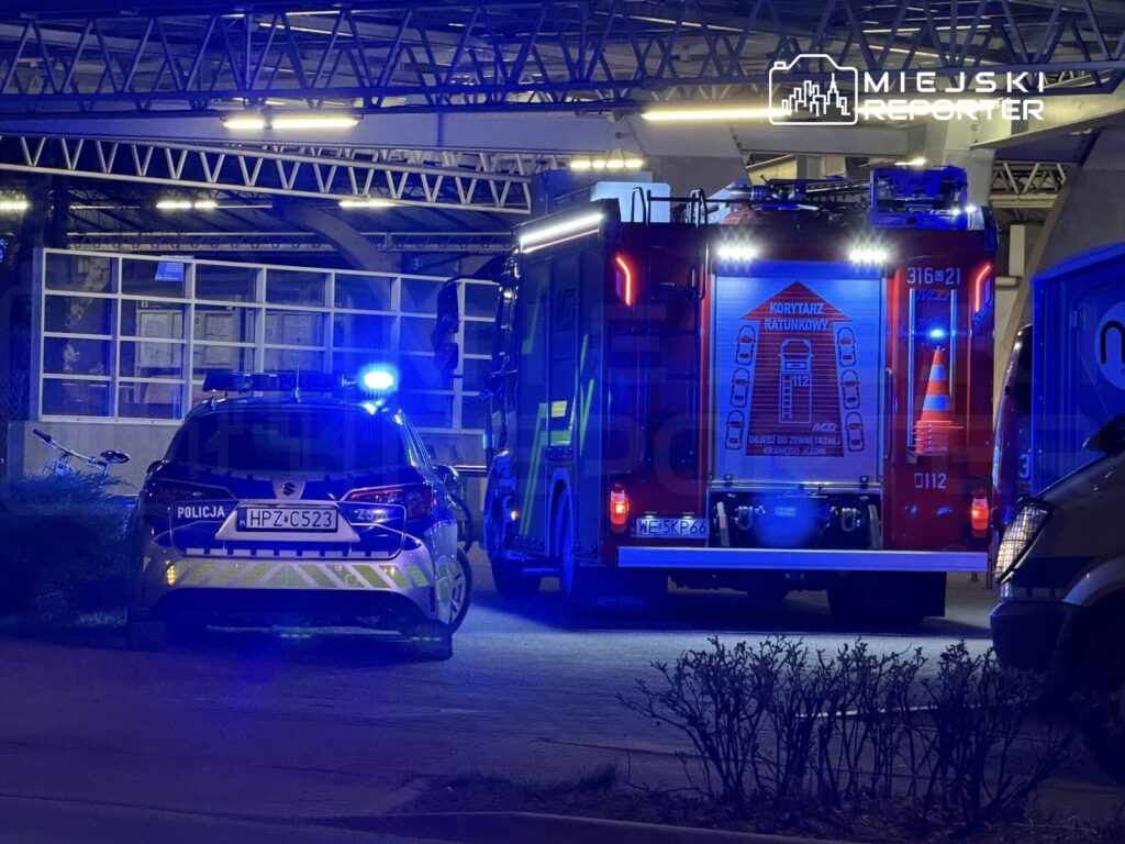 Radiowóz policyjny i wóz strażacki z włączonymi sygnałami świetlnymi na parkingu pod budynkiem.