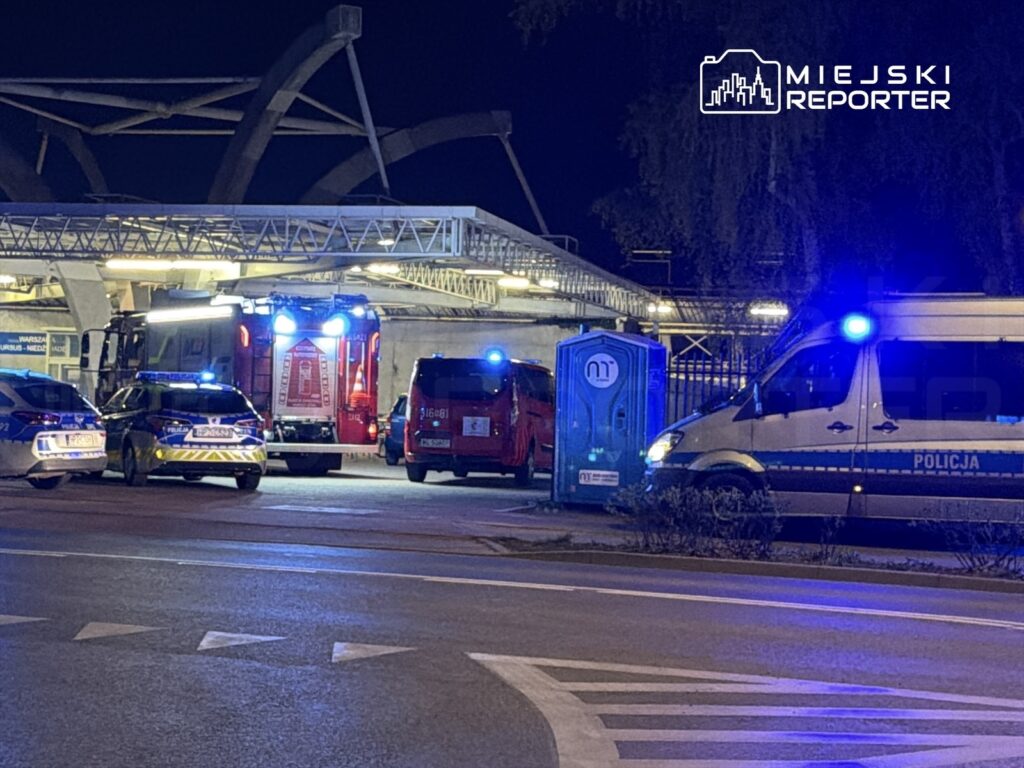W nocy pod nowoczesnym obiektem widać pojazdy służb ratunkowych i policji z włączonymi sygnałami świetlnymi.
