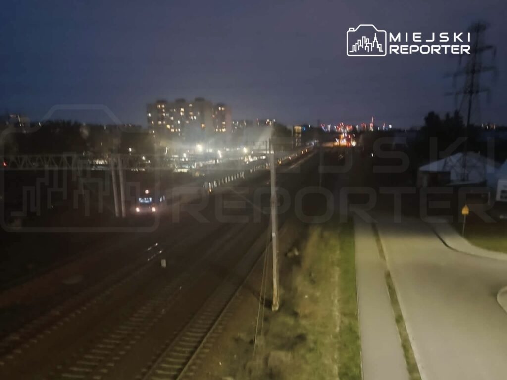 Pociąg przejeżdża po torach w oświetlonej okolicy, w tle widać budynki i miejskie światła nocą.