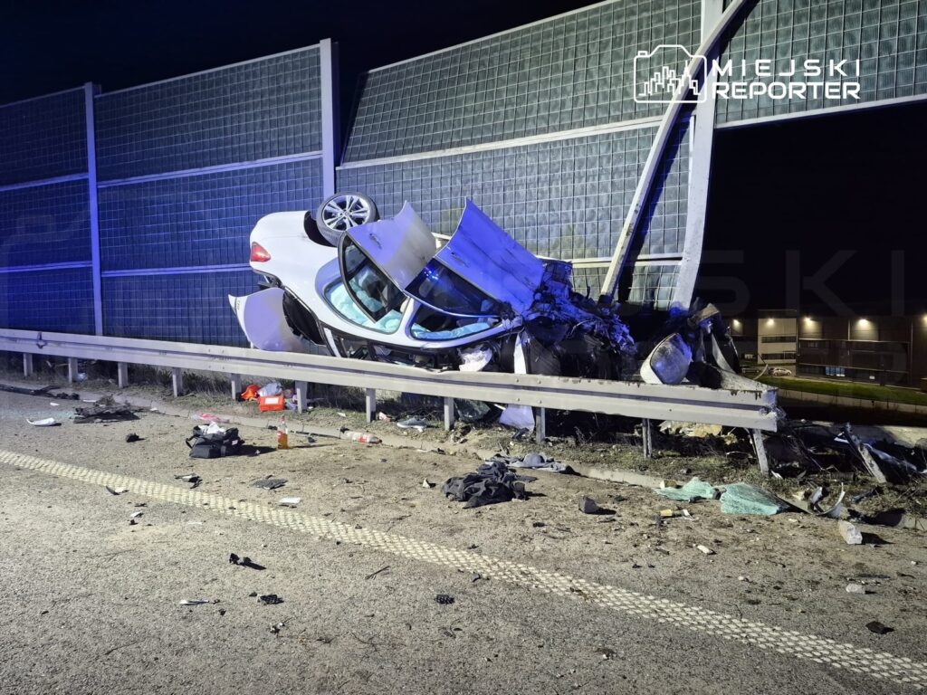 Zniszczony samochód osobowy leży na boku przy barierce, na drodze, wokół niego porozrzucane resztki oraz przedmioty.
