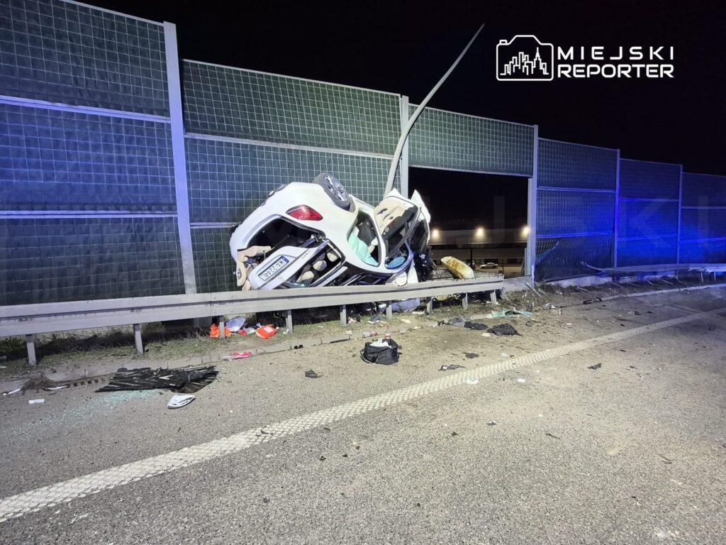 Rozbity samochód leży na barierce ochronnej przy drodze, otoczony odłamkami szkła i innymi szczątkami.