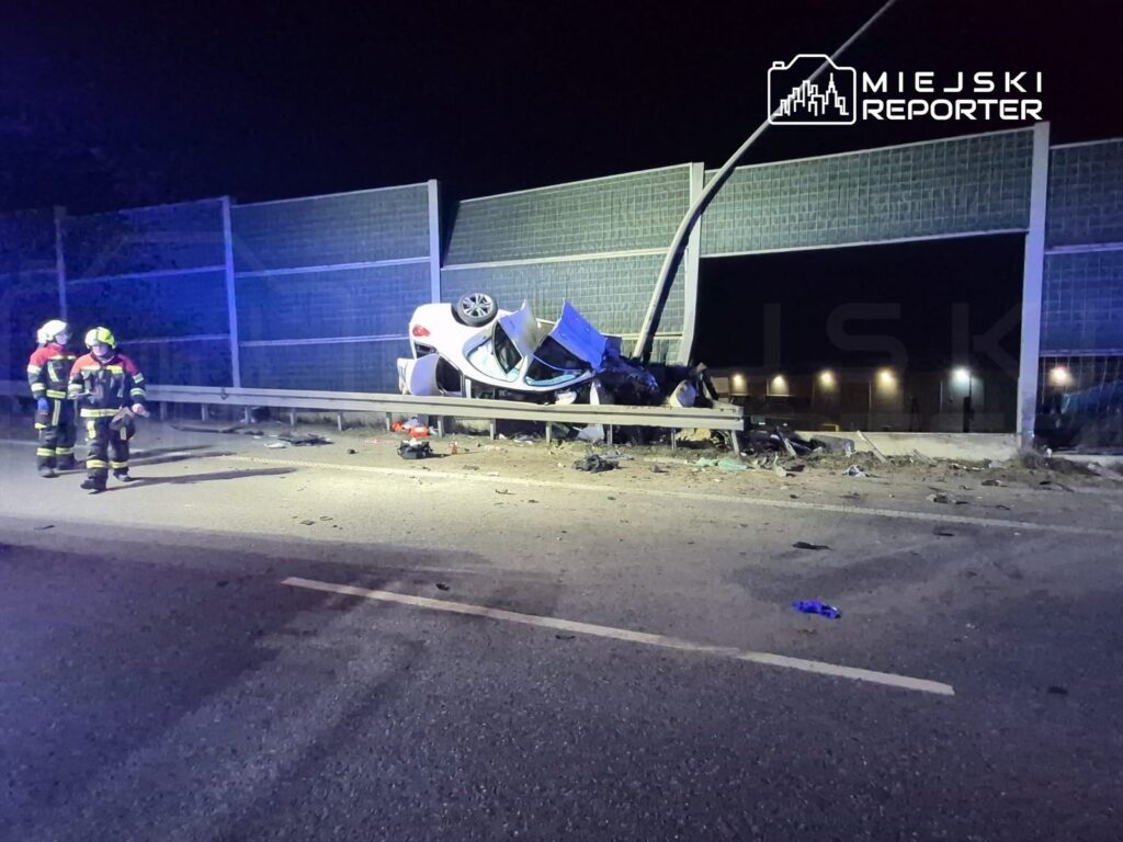 Dwa wozy strażackie i strażacy w mundurach zbliżają się do przewróconego samochodu na barierze drogowej w nocy.